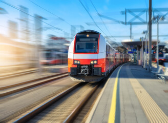 High-speed red passenger train moving at railway station platform under clear blue sky at sunset. Train station. Modern railway transportation concept with blurred motion effect. Railroad. Commercial