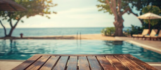 Fototapeta premium Blurry Background Image of Wooden Table at Poolside on Beautiful Beach Resort