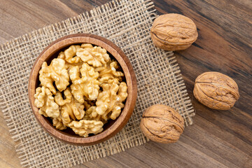 Overhead view of a bowl full of walnut kernels on a wooden surface. A piece of burlap and a few shelled walnut lying around