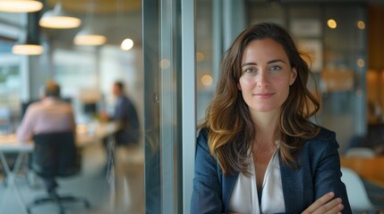 Obraz premium A poised female executive leans against a glass wall in a modern office, her gentle smile reflecting the vibrant energy of the bustling workspace behind her.