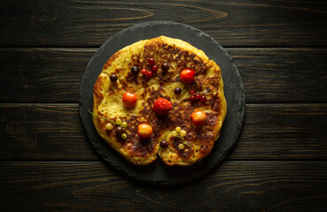 Round fried pancake with different ripe berries on black sorting board. Concept of serving tasty snack or dessert