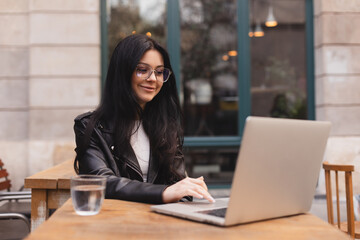 Student woman wear leather jacket, eyeglasses using laptop at cafe. Smiling young beautiful girl...