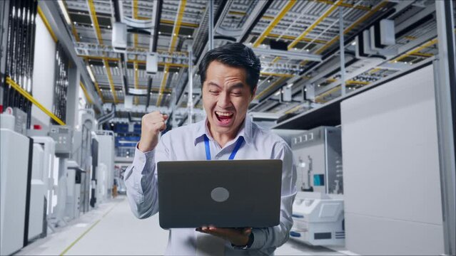 Asian Male Professional Worker Standing With His Laptop At Factory Digitalization, He Raises His Fist Up With Screaming Goal