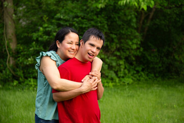 A mother and son with down syndrome embrace in a park. The woman is wearing a green T-shirt and the young man is wearing a red T-shirt. Both are smiling.
