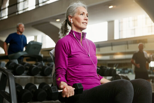 Dumbbell, music and wellness with mature woman on bench in gym for physical exercise or workout. Fitness, earphones and weightlifting with confident person in health club for active challenge