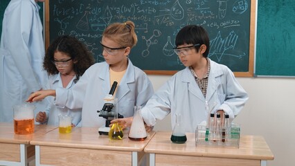 Boy pour solution in beaker while american girl inspect colored liquid. Energetic student support his friend doing experiment while teacher looking children explore and discover innovation. Pedagogy.