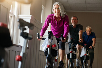 Woman, riding and bike at gym with earphone for fitness, training and workout listening to music for balance. Mature people, cycling machine and spin class exercise, wellness or cardio health goal