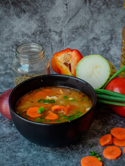 La imagen muestra un tazón de sopa con trozos visibles de verduras, como zanahorias y posiblemente algunas hojas verdes, sobre una superficie texturizada. Alrededor del tazón hay ingredientes.