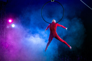 Professional aerial hoop woman gymnast circus performance at blue arena with dark background backlit with colored concert lights, performing gymnastic element © Michael Rekochinsky