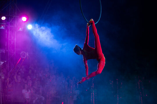 Professional aerial hoop woman gymnast circus performance at blue arena with dark background backlit with colored concert lights, performing gymnastic element