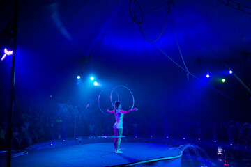 Young slim attractive circus gymnast woman performing live show with hula hoops on red arena with blue backlight, audience is excited