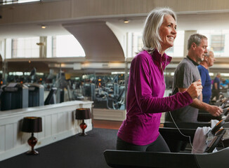 Fitness, cardio and mature woman on treadmill in gym for health, wellness and body exercise. Smile, earphones and female person listening to music on running equipment for workout in training center.