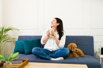 Joyful woman savoring a warm beverage while lounging on a couch with her adorable poodle in a cozy...