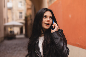 Happy brunette woman talking phone and look happy, smiling camera. Portrait of smiling female using mobile phone. Wow girl wear leather jacket. Female walk on the street.