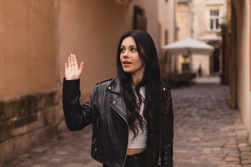 Fototapeta premium Happy attractive woman in black leather jacket, walk outdoors, meeting her friends or love, showing high five hey hello hi sign. She is joyful to meet her acquainted by chance. Girl wave her hand.