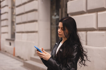 Outdoor shot of serious woman uses modern gadgets walks at street uses mobile phone, wear leather jacket sends sms tries to find route in unknown place in the city. Girl hold mobile phone and chatting