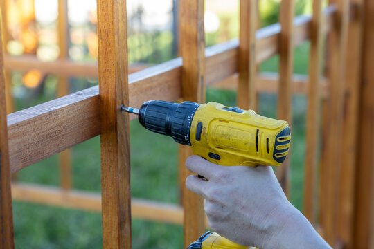 A person is using a drill to attach a wooden fence. The individual is focused on securing the fence panels to the posts using screws