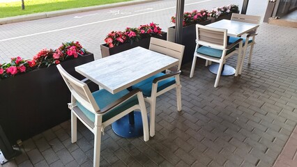 Wooden chairs and tables on metal bases stand on a tiled area. Nearby are tubs of ornamental flowers, and behind them is a sidewalk and bicycle path paved with concrete tiles