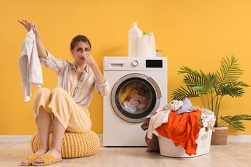 Young woman with smelly laundry sitting near washing machine on yellow background