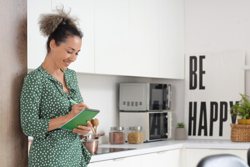 Beautiful middle-aged woman with notebook writing reminder in kitchen