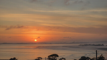 A burning sun rises and peers across a misty ocean bay through a muted sky.