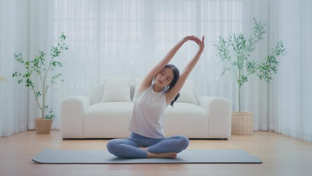 Happy young Asian woman practicing yoga and shoulder stretching at home sitting on floor in living room