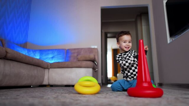 Lovely Caucasian toddler sits on the floor near the red conus. Cheerful baby puts a ring on the pyramid base. Low angle view.