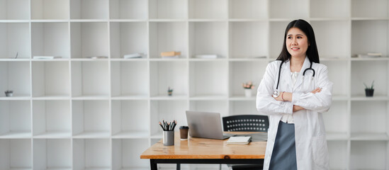 Confident female doctor in white lab coat with stethoscope, standing in modern office with laptop and bookshelves.