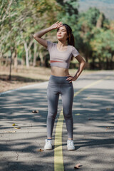 A woman is standing on a road, wearing a grey top and grey pants. She is looking up at the sky, possibly to check the weather