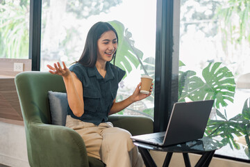 Naklejka premium A woman is sitting in a chair with a laptop open in front of her. She is holding a cup of coffee and she is smiling. The scene suggests a casual and relaxed atmosphere