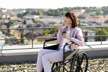 Smiling woman in wheelchair holding drink and smartphone on rooftop with scenic city view. Concept of accessibility, independence, and urban life.