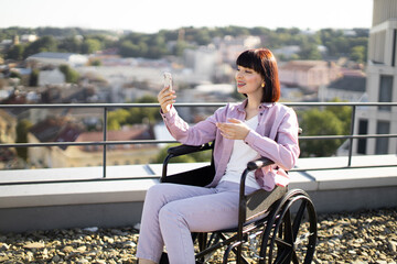 Young white woman in wheelchair enjoying video chat on smartphone while sitting on rooftop terrace overlooking city. She appears happy and content, with a beautiful urban landscape in the background.