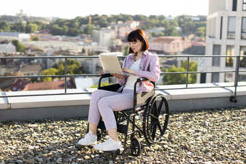 Positive Caucasian lady in wheelchair sitting with laptop typing message while working remotely. Business woman enjoys the view while working online on a roof top.