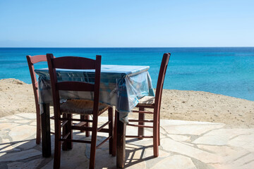 Open-air traditional seaside tavern in Greece. Empty wooden chair laid table on paved yard blue sea.