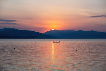 Colorful orange sky over sea water. Sun behind mountain silhouette, moored boat in open Greek sea.