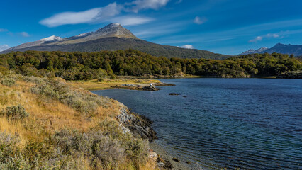 Obraz premium Beautiful autumn landscape of Patagonia. Yellowed grass and green forest grow on the shore of a blue lake. Ripples on the water. A mountain against an azure sky and clouds. Lapataia Bay. Argentina.