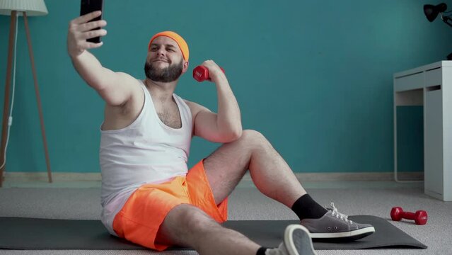 Full size man wear orange shorts spend time in home gym do selfie on mobile cell phone hold dumbbells sitting on floor