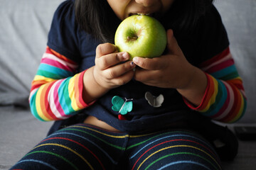 little girl bitting a green apple while sitting on a sofa 