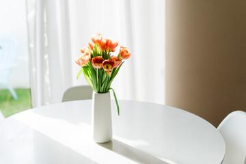Orange tulips in a white vase on a round table, bathed in sunlight.