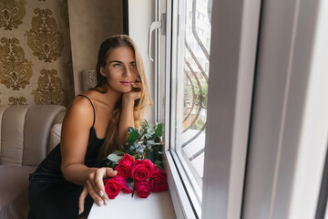 A woman in a black dress is sitting in front of a window with a bouquet of red roses.