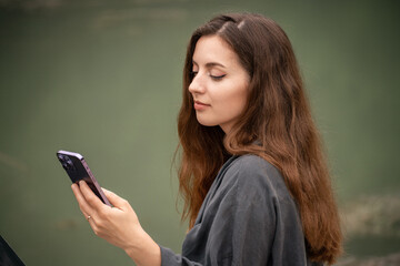 A woman is looking at her phone while sitting on a bench. She is wearing a black jacket and has long brown hair.