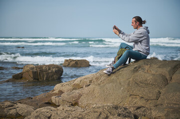Man sitting on rocky shoreline taking photo with smartphone at the beach