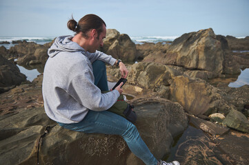 Man sitting on rocky coastline using smartphone by the ocean in casual attire