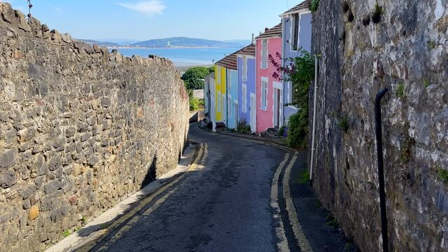 Walking down stone walled road with multi coloured houses in tourist resort, in south Wales, sea and beach seen in the background. It says fishing village, but it looks like seaside tourist resort. 