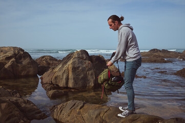A man in casual attire explores a rocky beach while carrying a backpack. The scene depicts a...