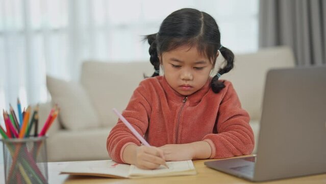 Asian young girl sitting at a desk with a laptop and a notebook. She is writing in the notebook and using a pencil. The scene suggests that she is working on a school assignment or studying for a test