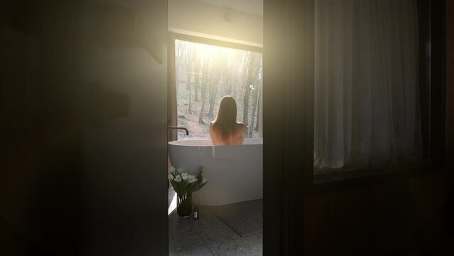 Woman enjoying bath with forest view. A woman relaxing in a bathtub with a view of the forest through a large window, bathed in warm sunlight.