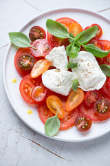 Plate of italian caprese with buffalo mozzarella, tomatoes and green basil leaves, vertical shot on a white granite surface, middle close-up