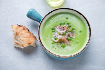 Turquoise serving bowl with green pea and bacon soup, horizontal shot on a light-grey granite surface, elevated view