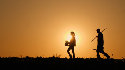 Against the field backdrop, silhouettes of two farmers walk with working equipment.